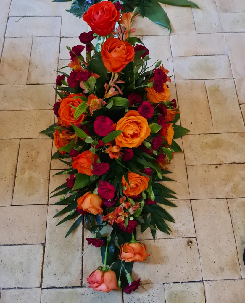 Blomsterdekoration med orange roser, mørkerøde nelliker og grønne blade som en smuk sidste hilsen ved begravelse eller bisættelse.